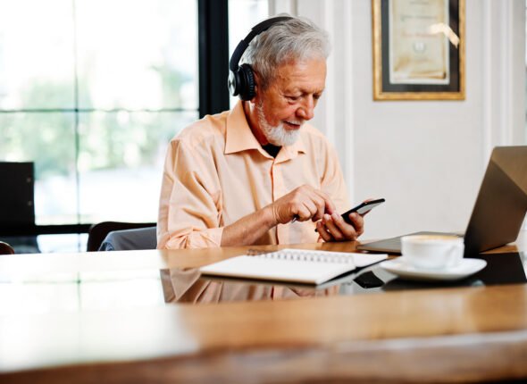 A dedicated senior student is sitting in a cafeteria with headphones on and listening online lecture on the laptop.
